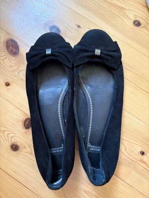 Svarta ballerinaskor med rosett - Stilrena svarta ballerinaskor i mockaimitation med en stor dekorativ rosett framtill och en liten metallisk detalj i mitten. Skorna har rundad tå och låg sula, perfekta för en trendig och enkel look. Säljer då de tyvärr är lite för stora för mig💕💕