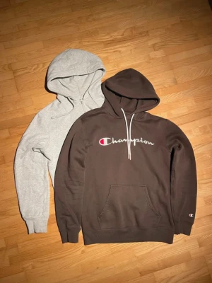 2-pack hoodies: Champion & grå zip - Säljer två hoodies: en brun Champion hoodie med broderad logga och känguruficka samt en ljusgrå zip-hoodie i medium men passar small bättre. Båda har långa ärmar och ribbade muddar. Perfekta för en avslappnad stil.