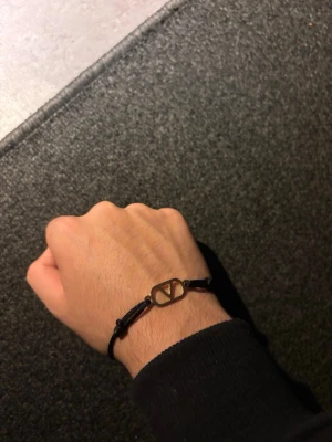 Valentio armband - Stilrent svart armband med justerbart band och en guldfärgad metallberlock i geometrisk form. Perfekt accessoar för dig som gillar minimalistisk och modern stil. Enkel att matcha med andra smycken och passar till många olika looks.
