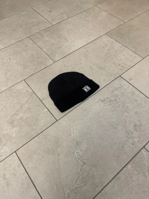Svart ribbad mössa med patch - Säljer en svart ribbstickad mössa med uppvikt kant och en snygg patch framtill. Klassisk och enkel design som passar till det mesta. Perfekt för kalla dagar när du vill hålla stilen.