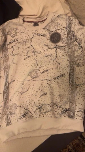 Vit sweatshirt med Versace-mönster - Unik vit sweatshirt från Versace med svart spräckligt mönster och klassiska Medusa-huvuden. Tröjan har tryckta Versace-loggor och dekorativa grekiska bårder längs sidorna. Rund halsringning och ribbade muddar. Perfekt för dig som vill sticka ut med en exklusiv look.