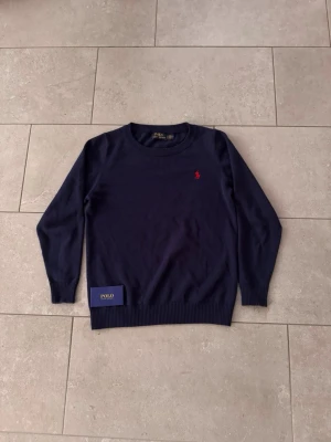Mörkblå tröja från Polo Ralph Lauren - Mörkblå rundhalsad tröja från Polo Ralph Lauren med den klassiska röda loggan broderad på bröstet. Tröjan har långa ärmar och ribbade muddar vid ärmslut och nederkant. Perfekt för en stilren och tidlös look.