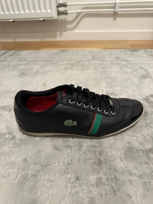 Svarta Lacoste sneakers med gröna detaljer - Snygga svarta sneakers från Lacoste med grönt och rött band på sidan samt den klassiska krokodilloggan. Skon har svarta snören, röd insida och är tillverkad i slätt lädermaterial. Perfekt för dig som vill ha en stilren och sportig look.