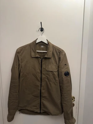 Beige overshirt med zip och ficka - Snygg beige overshirt med dragkedja framtill, klassisk krage och en bröstficka med lock. Ärmen har en rund svart detalj som ger en cool touch. Tillverkad i ett tunt bomullsliknande material, perfekt för lager på lager.
