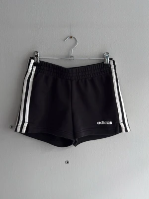Svarta Adidas shorts med vita ränder - Klassiska svarta shorts från Adidas med vita ränder längs sidorna och Adidas-logga på ena benet. Elastisk midja för skön passform. Perfekta för träning eller chill dagar.