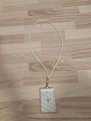 Vit korthållare med snodd - Snygg vit korthållare med klassiskt mönster och blå logga framtill. Kommer med matchande vit snodd och guldfärgad metallklämma. Perfekt för att bära kort runt halsen på ett stilrent sätt.