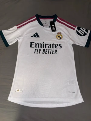 Real Madrid matchtröja Adidas - Snygg vit Real Madrid matchtröja från Adidas med klubbmärke på bröstet och sponsortryck. Tröjan har mörkgröna detaljer vid krage och ärmslut samt vinröda ränder på axlarna. Tillverkad i lätt och ventilerande material, perfekt för fotbollsträning eller match.