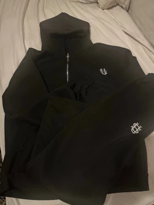Svart loungewear set med brodyr - Mysigt svart loungewear set med hoodie och byxor. Hoodien har dragkedja framtill och båda plaggen har en vit broderad logga. Setet är gjort i mjukt material och har en avslappnad passform, perfekt för chill dagar hemma.