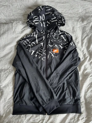Nike svart vindjacka med bokstavsmönster - Snygg svart vindjacka från Nike med vitt bokstavsmönster över hela huvan och axlarna. Jackan har dragkedja framtill, två fickor med dragkedja och en patch med 'Just Do It.' och Nike-loggan på bröstet. Tillverkad i lätt och vindtätt material.