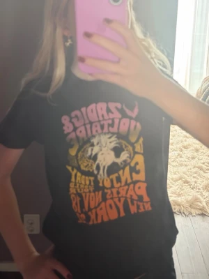 Svart Zadig & Voltaire t-shirt - Cool svart t-shirt från Zadig & Voltaire med färgglatt tryck i rosa, gult och orange framtill. Trycket har en retro tour vibe med text om Paris och New York samt en grafisk illustration av en häst. Perfekt för dig som gillar statement-plagg.