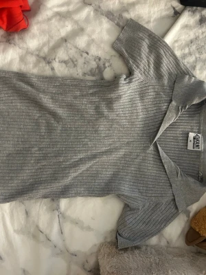 Grå ribbad polotopp med krage - Säljer en grå ribbad polotopp med korta ärmar och bred krage. Toppen har en slim passform och är tillverkad i ett mjukt, stretchigt material som sitter snyggt på kroppen. Perfekt att styla med jeans eller kjol för en clean look.