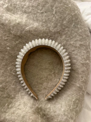 Vitt diadem med pärlbroderi - Säljer ett vitt diadem dekorerat med glittrande pärlor i flera rader. Diademet har en bred form och insidan är klädd i beige tyg för extra komfort. Perfekt accessoar för att lyfta din look med en elegant touch.