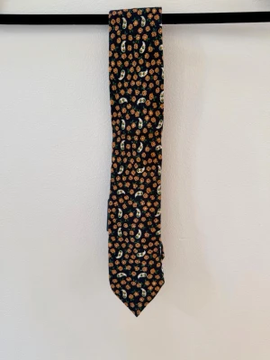 Breuer – Exklusiv slips i 100 % ull - Elegant och exklusiv Breuer-slips från det anrika franska herrmodemärket Breuer, grundat 1892.  Slipsen har ett stilfullt, tätt mönster med orange blommor och vita paisley-liknande detaljer mot en mörk marinblå bakgrund. En sofistikerad och tidlös design som passar perfekt till kostym och dressad klädsel.  Tillverkad i 100 % ull med en exklusiv vävd kvalitet och ett elegant fall.  ✨ Helt ny – prislappen sitter kvar 💰 Ordinarie pris: 995 kr 🧵 100 % ull 🇫🇷 Breuer – anrikt franskt herrmode sedan 189