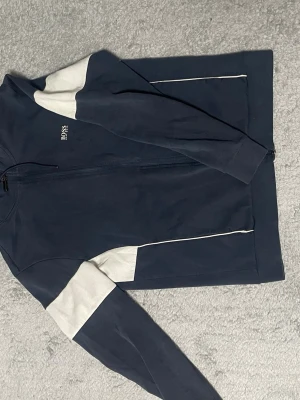 Mörkblå hoodie med vita detaljer BOSS - Snygg mörkblå hoodie från BOSS med dragkedja framtill och vita paneler på ärmarna. Liten BOSS-logga broderad på bröstet. Hoodien har en sportig look och är tillverkad i mjukt material, perfekt för en avslappnad stil.