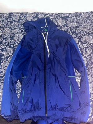 Blå vindjacka från Lacoste Sport - Snygg blå vindjacka från Lacoste Sport med huva och dragkedja framtill. Jackan har gröna detaljer vid fickorna och meshfoder på insidan. Perfekt för blåsiga dagar och sportiga outfits. Materialet är lätt och vattenavvisande.