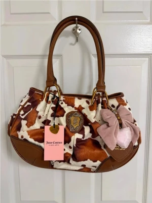Juicy Couture handväska med ko-mönster - Juicy Couture handväska i brunt och vitt ko-mönster med detaljer i brunt konstläder. Väskan har två handtag, en stor rosa rosett och ett emblem framtill. Söt och unik design med exklusiv tag kvar. Perfekt för dig som vill sticka ut!