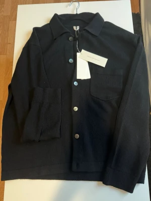 Cardigan arket - Stilren svart overshirt med klassisk krage och knappstängning framtill. Modellen har en bröstficka på vänster sida och är tillverkad i ett mjukt, stickat material. Perfekt att bära över en t-shirt för en avslappnad look. Helt ny!