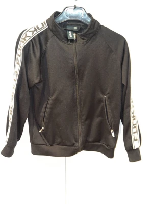 Svart zip-jacka från Funky - Svart sportig zip-jacka från Funky med vita detaljer och logga längs ärmarna. Jackan har två fickor med dragkedja och ribbade muddar vid ärmslut och nederkant. Perfekt för träning eller chill. Materialet är mjukt och stretchigt.