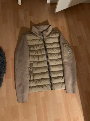 Beige dunjacka  - Snygg beige dunjacka med quiltad kropp och stickade ärmar. Jackan har hög krage, dragkedja framtill och en ficka. Perfekt för kalla vinterdagar och riktigt trendig look.
