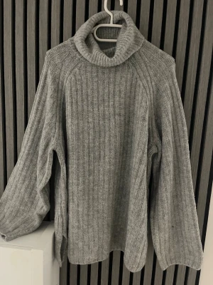 Grå polotröja från 157 - Mysig grå stickad polotröja från 157 med bred ribbstickning och hög krage. Tröjan har avslappnad passform och långa ärmar, perfekt för kalla dagar. Enkel att matcha med jeans eller kjol för en chill look.