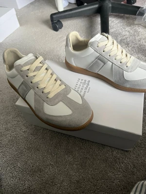 Maison margiela - Säljer ett par stilrena sneakers i vitt och ljusgrått med beige skosnören och gummisula. Skorna har paneler i mocka och slät läderimitation, samt klassisk låg profil. Perfekta för dig som gillar minimalistisk och clean stil.