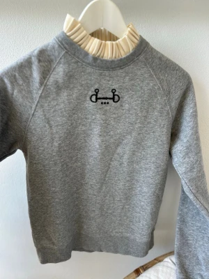 Ridtröja CRW xs Grå sweatshirt med broderad detalj - Mysig grå sweatshirt med svart broderad detalj på bröstet. Tröjan har rund halsringning, långa ärmar och ribbade muddar vid ärmslut och nederkant. Perfekt för kalla dagar och enkel att matcha med andra plagg.