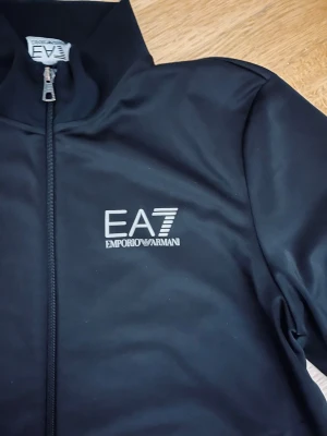 Svart jacka från Emporio Armani EA7 - Snygg svart jacka från Emporio Armani EA7 med hög krage och dragkedja.  Perfekt för dig som gillar stilrena plagg. Den använd jättelitet och är som ny.