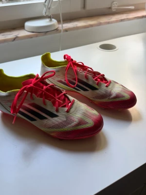 Vita och rosa fotbollsskor Adidas - Snygga fotbollsskor från Adidas i vitt med rosa och neongula detaljer samt svarta ränder på sidorna. Skorna har rosa snörning och låg profil, perfekta för snabba vändningar på planen. Ovandelen är i lätt syntetmaterial för bra bollkontroll.