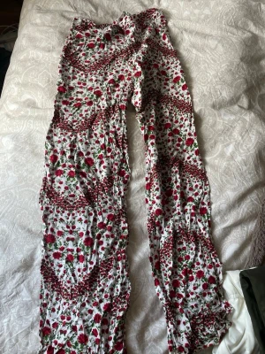 Blommiga vida leggings i vitt och rött - Säljer ett par supervåfflade leggings med blommigt mönster i rött, grönt och vitt. De har en lös och flowy passform med resår i midjan och är gjorda i ett tunt, mjukt tyg som känns riktigt skönt mot huden. Perfekta för dig som gillar boho-vibe och färgglada plagg!