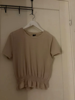 Beige topp med rynkad midja - Söt beige topp med korta ärmar och rund halsringning. Toppen har en rynkad midja med volangkant nedtill som ger en feminin touch. Perfekt till jeans eller kjol för en avslappnad men stilsäker look.