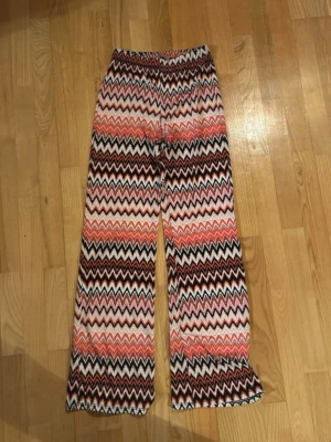 Mönstrade vida leggings i retrostil - Säljer ett par färgglada leggings med zigzag-mönster i rosa, orange, brunt och vitt. De har hög midja, resår i midjan och vida ben. Perfekta för dig som gillar retrovibbar och vill sticka ut med din stil!