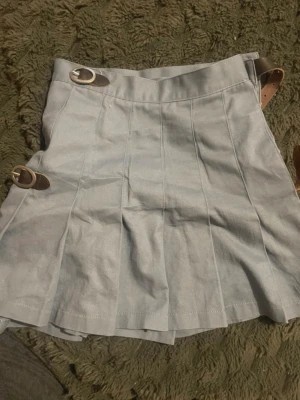 Brandy melville ljusblå kortkjol med spännen - Snygg ljusblå kortkjol med två dekorativa bruna spännen i midjan och på sidan. Kjolen har veckad design och en stilren look som passar till många outfits. Perfekt för dig som gillar en trendig och enkel stil. Man kan fixa storleken.