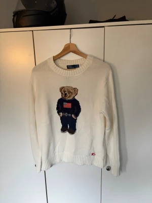 Vit stickad tröja med björn från Polo Ralph Lauren - Vit stickad tröja med rund hals och ikonisk björn på bröstet, klädd i mörkblå tröja med amerikansk flagga och jeans. RL-broderi vid nederkanten. Perfekt för dig som gillar klassisk och lekfull stil.