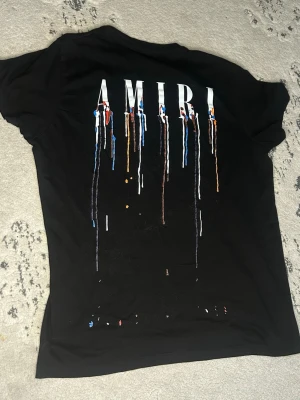  AMIRI  - Säljer en svart t-shirt från AMIRI med tryck i vitt och färgstänk i blått, rött, orange och gult både fram och bak. T-shirten har rund hals och korta ärmar. Perfekt för dig som gillar streetwear och vill sticka ut med en unik design. storlek M men skulle säga den sitter lika bra för S också 