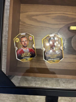 Fotbollssamlarbilder Topps - Två exklusiva fotbollssamlarbilder från Topps med Harry Kane och Jude Bellingham. Kortens design är guldfärgad med spelarnas porträtt och titlarna 'King of Headers' och 'Last-Minute Hero'. Perfekt för dig som gillar fotboll och samlarobjekt.