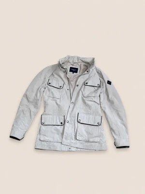 Hackett London - Stilren vit filed jacket från Hackett London med fyra framfickor och svarta detaljer. Jackan har hög krage, tryckknappar och patch på ärmen. Perfekt för dig som gillar klassisk och clean stil.