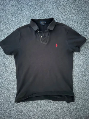 Svart pikétröja från Polo Ralph Lauren - Klassisk svart pikétröja från Polo Ralph Lauren med röd broderad logga på bröstet. Tröjan har korta ärmar, krage och knappar vid halsen. Perfekt till både jeans och shorts för en clean och stilren look.