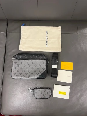 Crossbody väska    - Snygg handväska från i svart och grått med klassiskt monogrammönster. Väskan har en rektangulär form, avtagbar axelrem och matchande liten pouch. Tillverkad i canvas med läderdetaljer. Perfekt för dig som gillar exklusiva accessoarer.