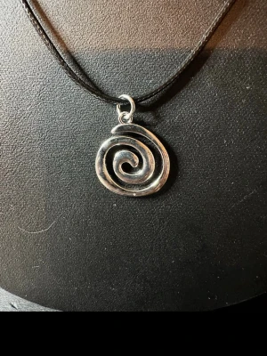 Halsband med spiralformad silverberlock - Snyggt halsband med en spiralformad berlock i silverfärgad metall. Hängs på en tunn svart rem som ger en stilren och modern känsla. Perfekt för dig som gillar unika detaljer och enkel design.