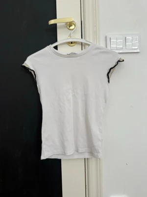 Vit topp med svart guldiga detaljer - Säljer en vit topp med korta ärmar och svart guldiga detaljer längs ärmsluten. Toppen har en enkel, figurnära passform och är tillverkad i mjukt bomullsmaterial. Perfekt för dig som gillar minimalistisk stil med en twist.