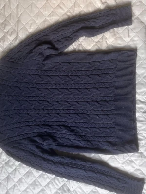 Mörkblå kabelstickad tröja - Mysig mörkblå kabelstickad tröja med långa ärmar och ribbade muddar vid ärmslut och nederkant. Klassisk design med flätmönster över hela tröjan. Perfekt för kyliga dagar och enkel att matcha till olika outfits.