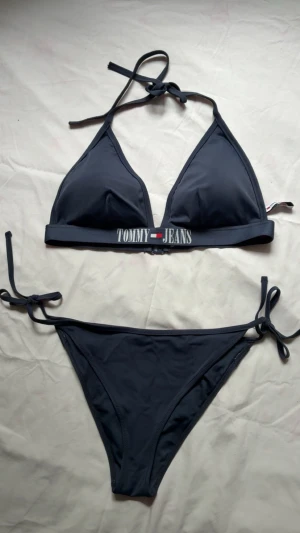 Tommy Jeans triangel bikini set - Mörkblått Tommy jeans bikini set. Köpte under och överdelen separat för 3 år sedan för ca 900 kr men nu är de alldeles för stora för mig. Har knappt används då jag inte badade så mycket förut. Överdelen är i storlek XL och axelbanden kan knytas i ryggbandet om man föredrar det då det finns öglor där (se bild 2). Underdelen är i storlek L. Kan säljas separat om det finns intresse. Jag hade kanske BH storlek 80G eller 80F när jag köpte överdelen men kan mäta ryggbandets exakta storlek.