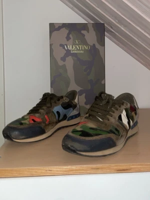 Valentino Garavani Rockrunner Camouflage strl 42 + Box - Valentino Garavani Rockrunner sneakers i camouflage, storlek 42.  Skorna är använda och har bruksslitage enligt bilderna. Det finns en mindre spricka/slitage framtill i tyget, men den går inte igenom. Sulan har även börjat släppa lite längst fram, vilket syns på bilderna.  I övrigt är skorna hela och användbara. Original Valentino Garavani-box medföljer.  Se bilderna noggrant för exakt skick. Fler bilder kan skickas vid intresse.