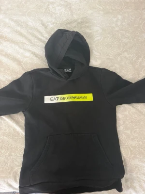 Svart EA7 Emporio Armani hoodie - Snygg svart hoodie från EA7 Emporio Armani med stor huva och magficka. På bröstet finns en vit och neongul logga med texten EA7 Emporio Armani. Perfekt för en avslappnad och trendig stil. Köpte i jd för 1200kr och den är s eller m
