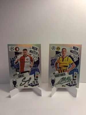Match Attax fotbollskort Man of the Match - Två Match Attax fotbollskort med Man of the Match-tema. Ena kortet visar Gimenez från Feyenoord i rödvit tröja och det andra Schlotterbeck från Borussia Dortmund i gulsvart tröja. Båda korten har holografisk effekt och cool design.