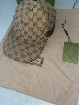 Beige keps från Gucci med GG-mönster - Snygg beige keps från Gucci med det klassiska GG-monogrammönstret över hela kepsen. På sidan finns en grön och röd rand som ger en extra lyxig touch. Tillverkad i canvasmaterial och levereras med påse, box och tagg.