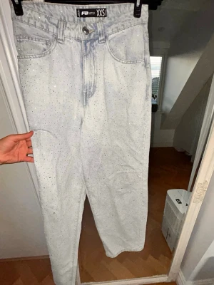 Ljusblå jeans med strassdetaljer - Snygga ljusblå jeans från FST SSTR i storlek XXS. Jeansen har raka ben och är dekorerade med små strassdetaljer över hela tyget för en extra glittrig look. Klassisk femficksmodell och hög midja. Perfekta för dig som vill sticka ut lite extra.