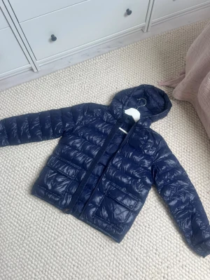 Mörkblå dunjacka från Polo Ralph Lauren - Snygg mörkblå dunjacka från Polo Ralph Lauren med huva och två stora fickor framtill. Jackan har en glansig finish och quiltad design som ger en modern look. Det står XL (16) på prislappen, men den passar mig som har XS-S. 