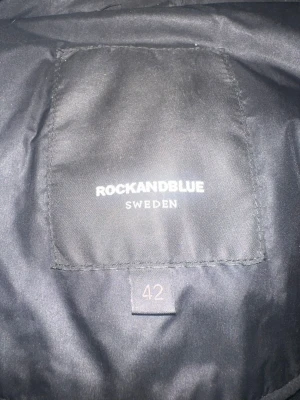Svart lång dunjacka Rockandblue - Snygg svart lång dunjacka från Rockandblue med stor fluffig äkta päls på huvan och quiltade detaljer. Jackan har dragkedjor och knappar framtill, samt flera fickor med dragkedja. Perfekt för kalla dagar och riktigt varm.