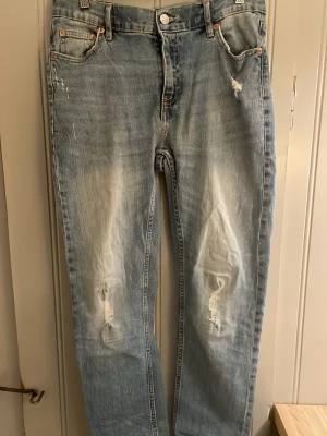 Ljusblå slitna raka jeans - Säljer ett par ljusblå jeans med raka ben och slitningar på båda knäna. Jeansen har klassisk femficksmodell och är tillverkade i denim. Perfekta för en avslappnad och trendig look.